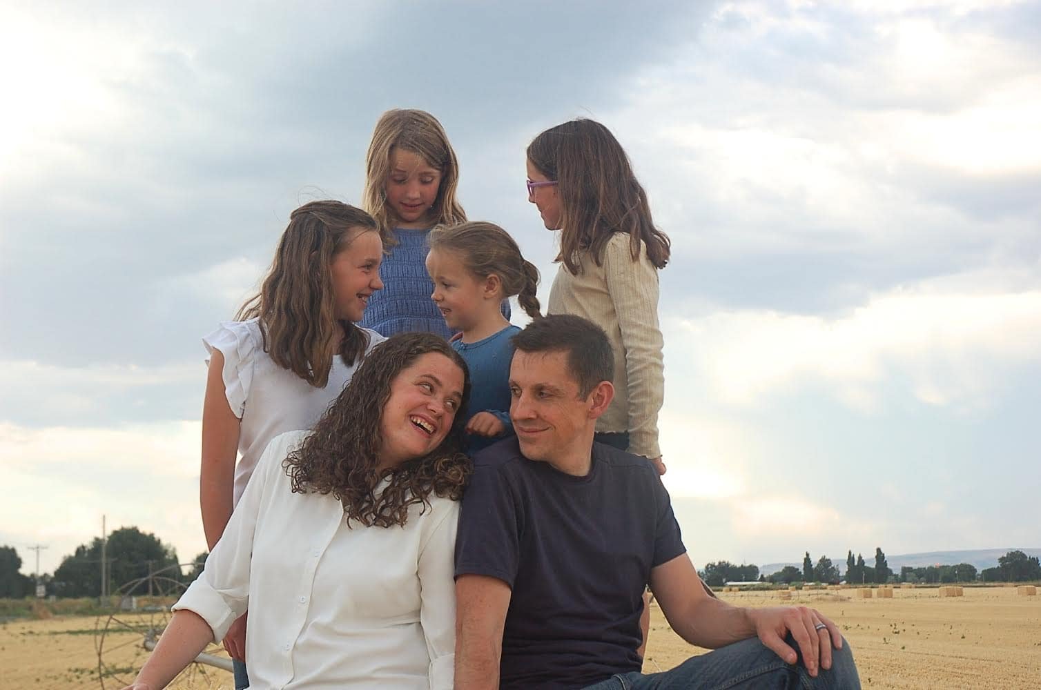 Picture of family (mom, dad, 4 girls) outside sitting on a hay bale in the middle of a field.  Everyone is looking at each other and smiling.